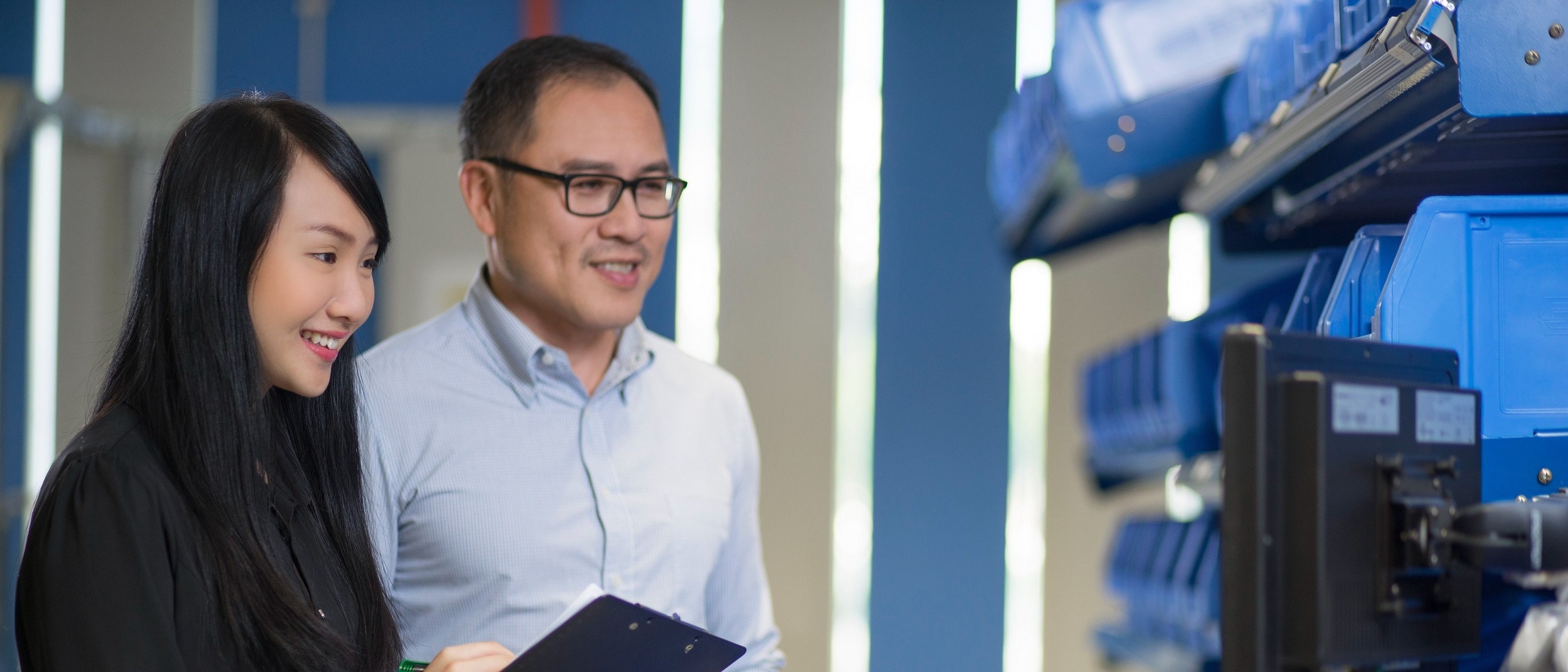 A woman and a man smile while reviewing a document in a warehouse, surrounded by blue storage bins